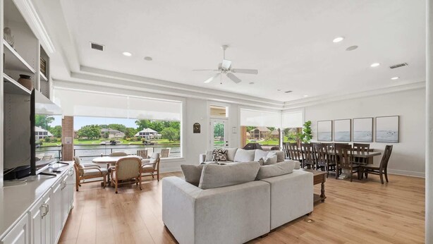 A spacious living area with a large gray sofa, dining table, and chairs, a kitchen with white cabinets, and a view of a residential area. - Lakefront House with Huge Swim Spa (Montgomery)
