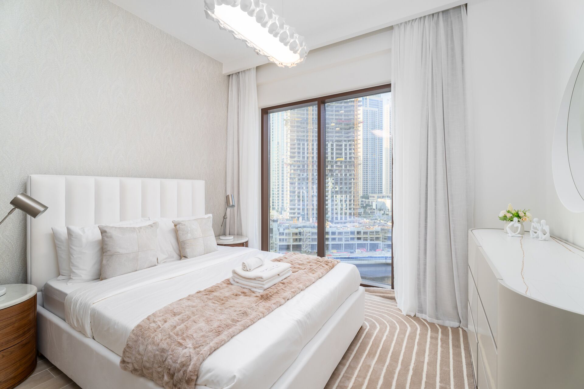 Ein modernes Hotelzimmer mit einem großen Bett, einem Nachttisch, einem Schminktisch mit Spiegel und Blick auf hohe Gebäude durch das Fenster.