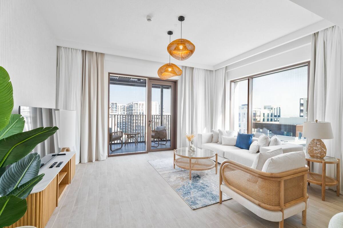 A modern living room with a sofa, coffee table, and rattan chair. There is a balcony with a view of the city.