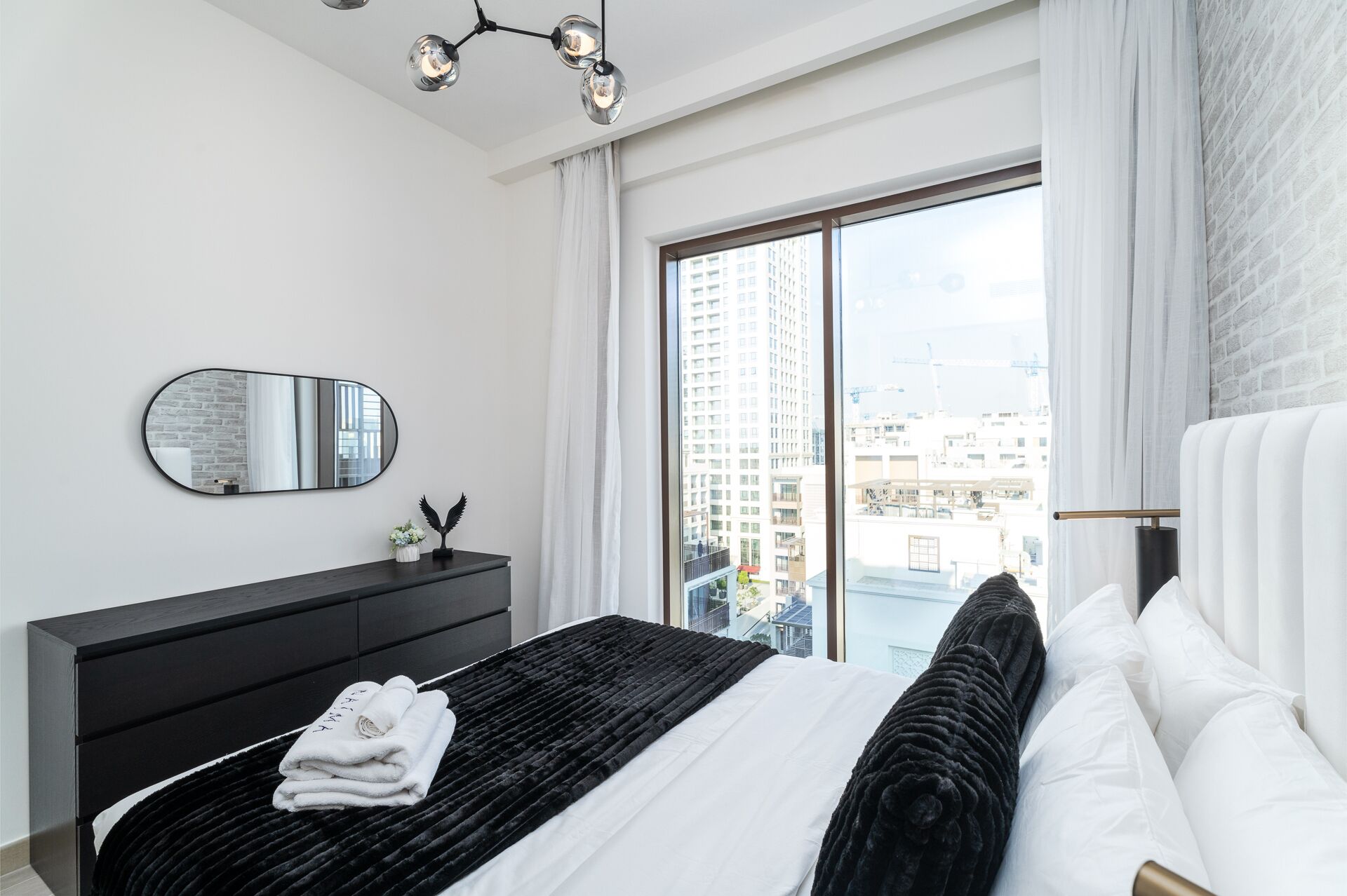 Un dormitorio moderno con una cama grande, un aparador negro, un espejo redondo y vistas de edificios altos a través de un ventanal.