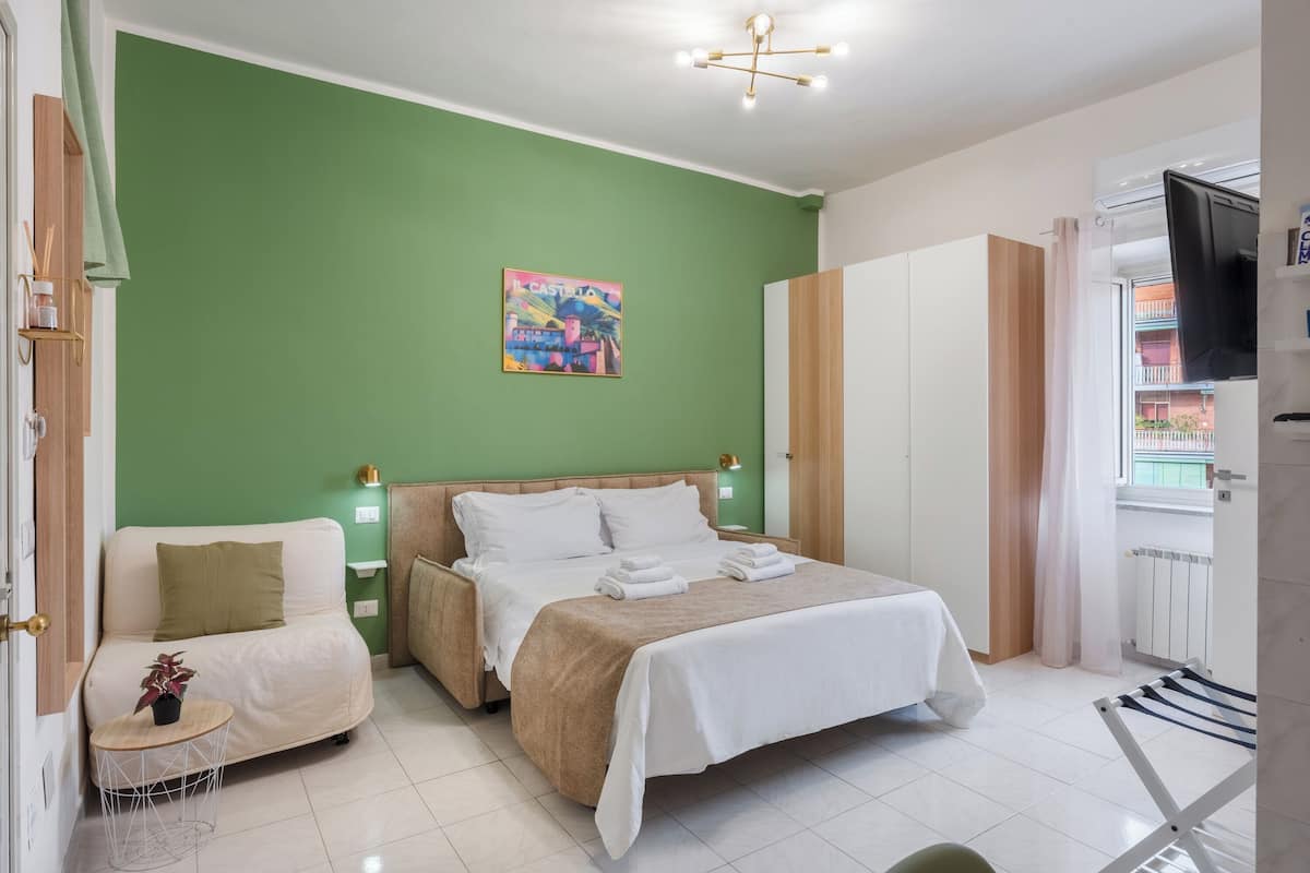 Una habitación de hotel con pared verde, una cama con ropa de cama blanca, un sofá, una mesita con una planta y un televisor montado en la pared.