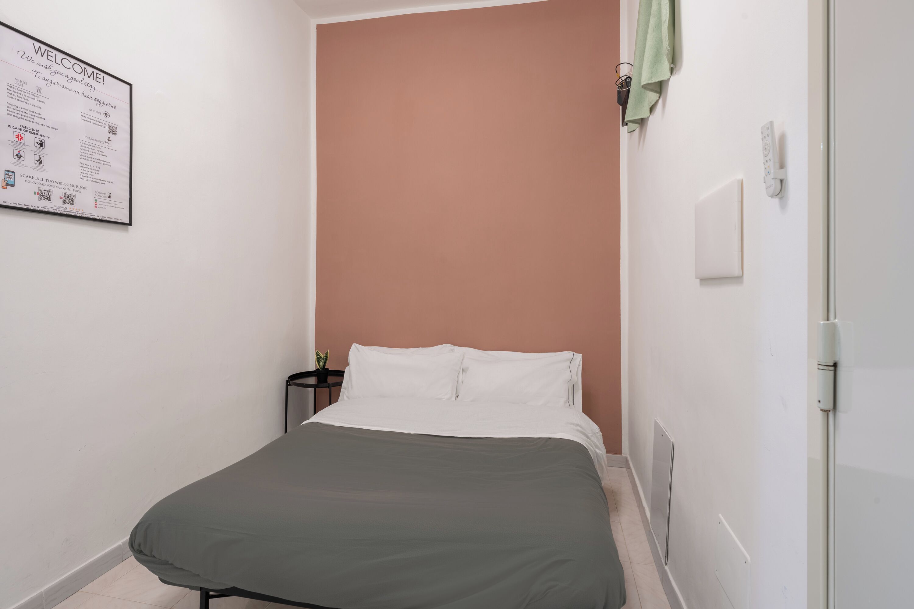 Un dormitorio pequeño y ordenado con cama, luz en la pared y un cartel de bienvenida.