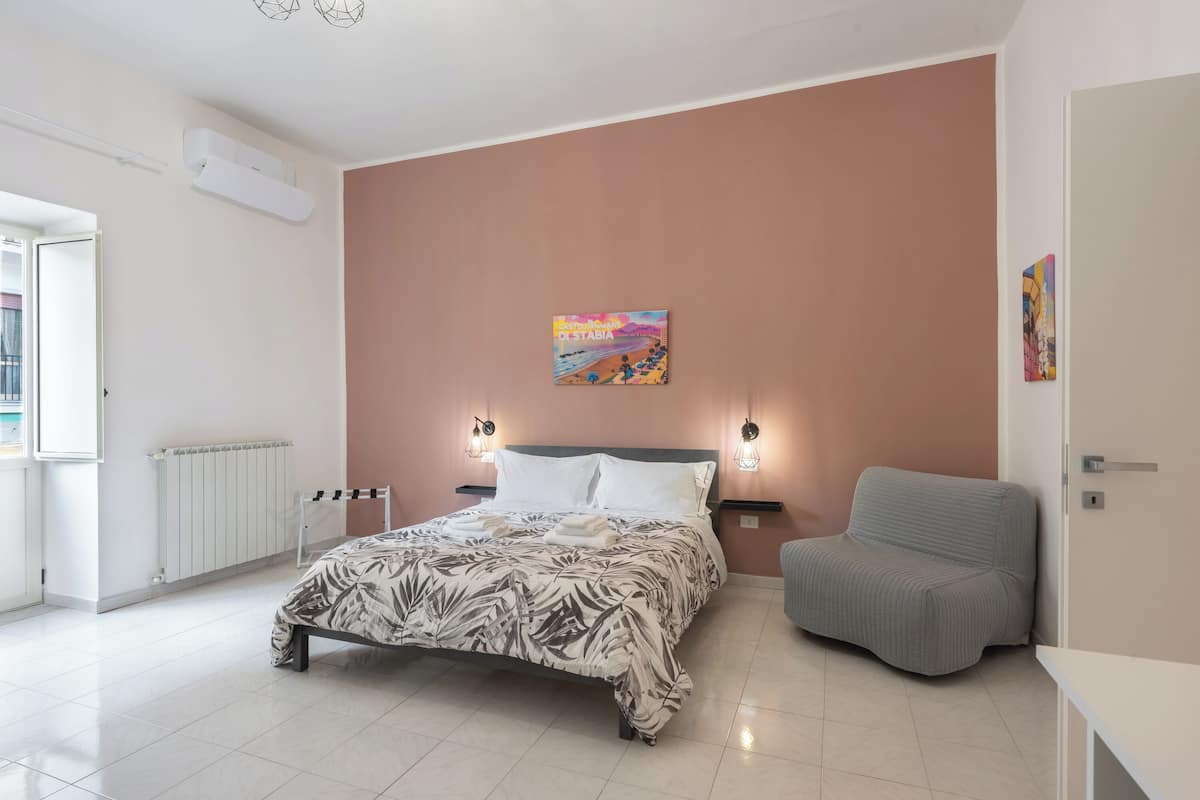 Un dormitorio con cama, un sillón gris, un radiador y una obra de arte colgada en la pared.