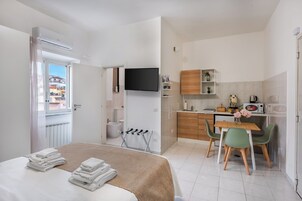 Una habitación de hotel moderna con una cama, una pequeña kitchenette, un área de comedor con mesa y sillas, un televisor montado en la pared y un baño visible a través de una puerta abierta.