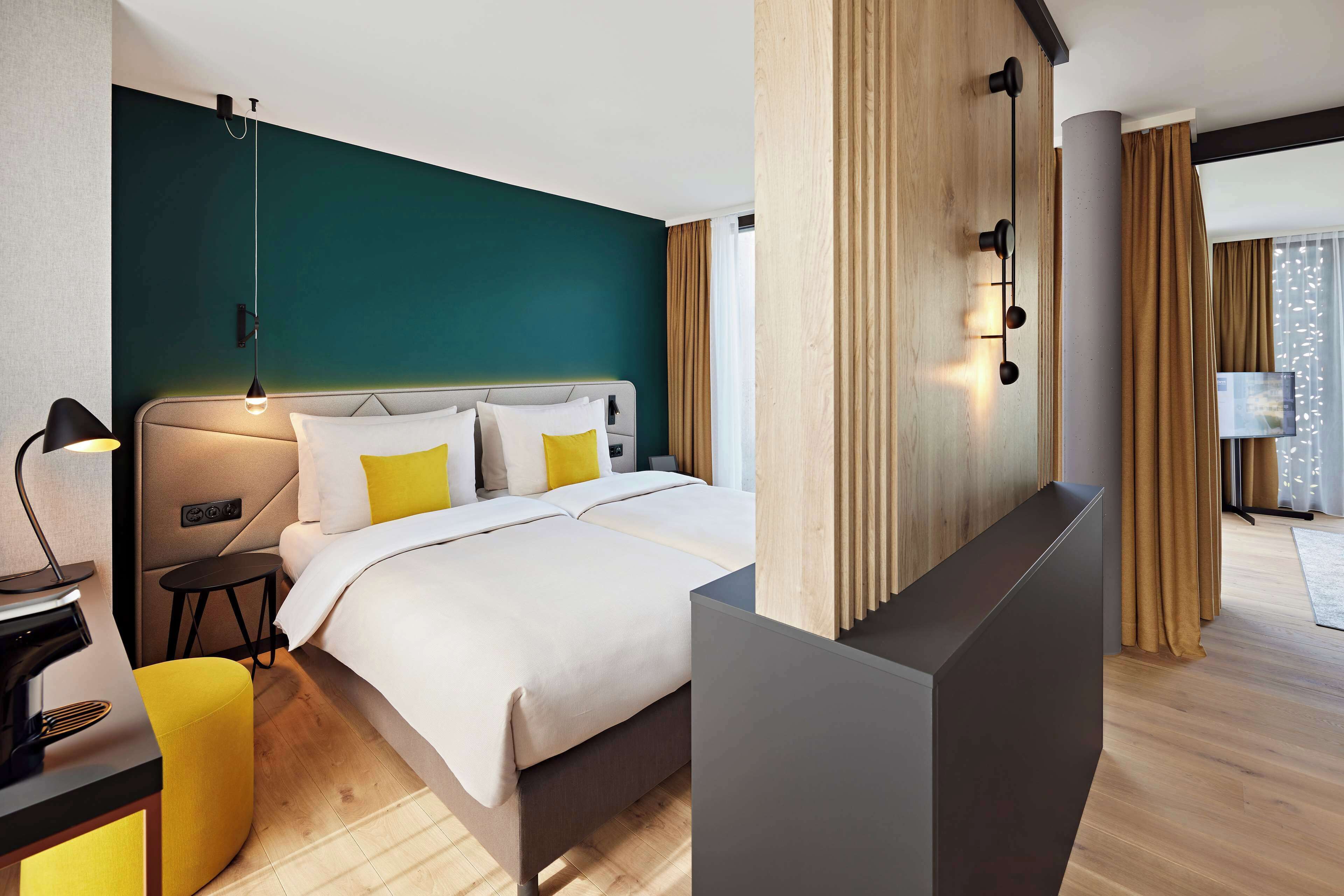 Een moderne hotelkamer met een bed, nachtkastje, lamp en uitzicht op de stad door grote ramen.