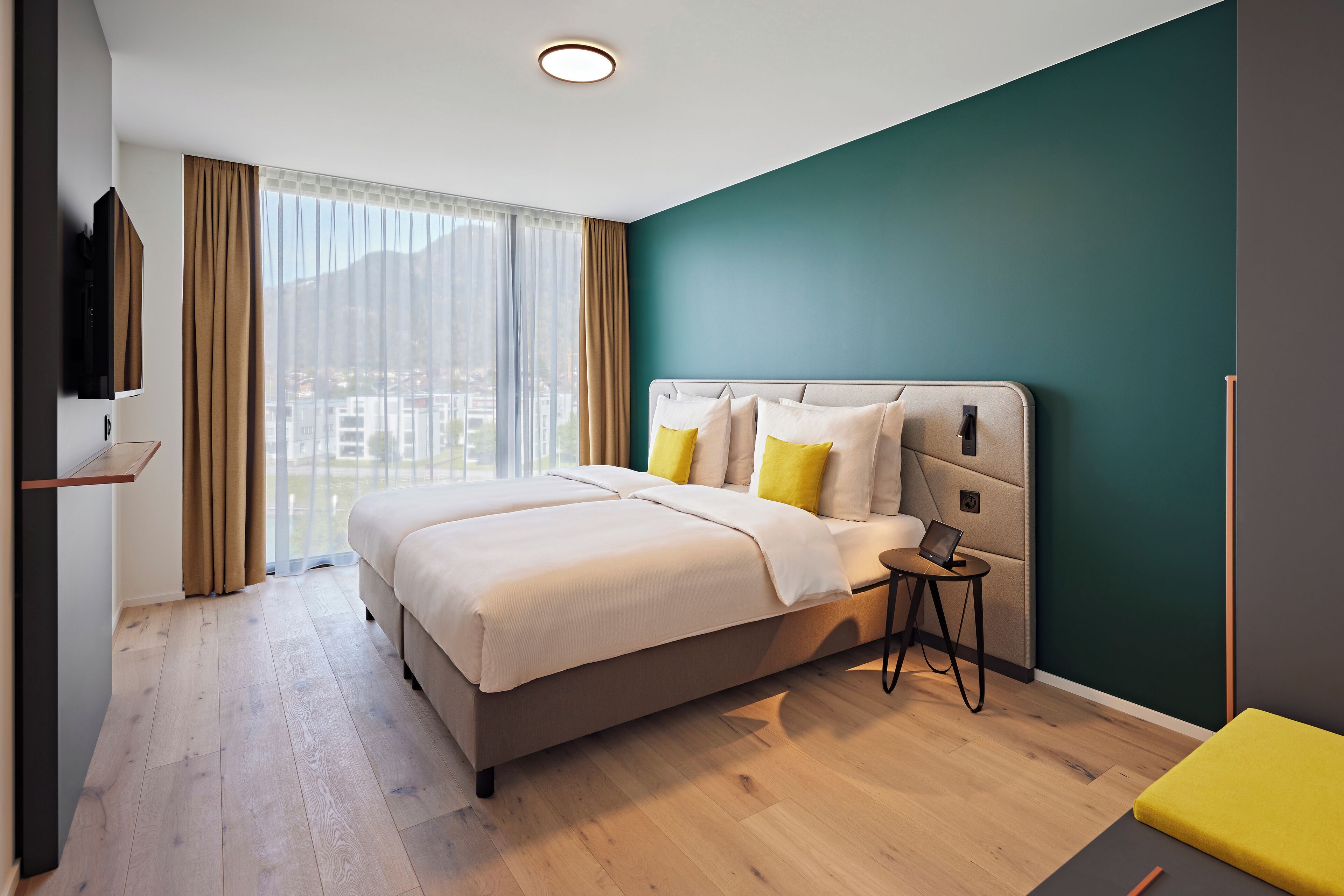 A modern hotel room with a large bed, a bedside table with a lamp, and a view of the city through large windows.