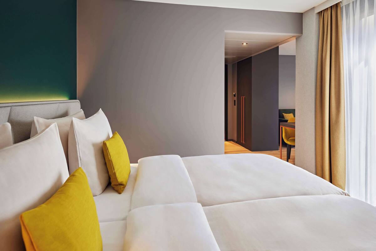 A modern hotel room with a large bed, a headboard, and a view of another room through an open door.