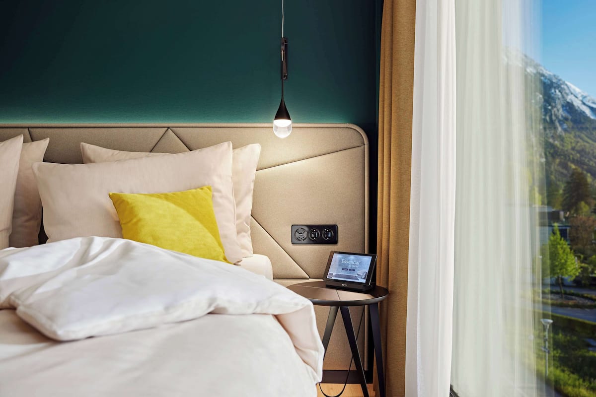 A hotel room with a bed, a bedside table with a laptop, a wall-mounted control panel, and a view of a mountain through a window.
