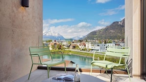 Ein Balkon mit zwei grünen Metallstühlen, einem Tisch mit Flasche und Gläsern und Blick auf einen Berg sowie Gebäude über einem Gewässer.