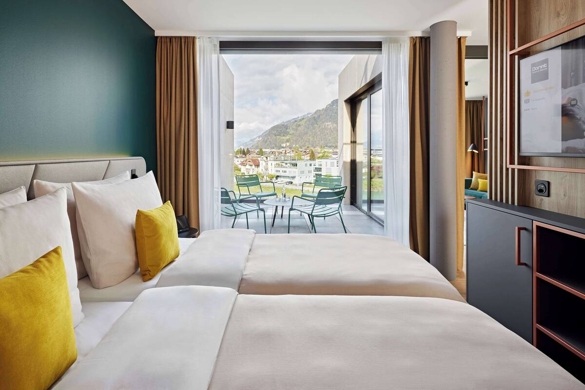 A hotel room with a bed, a TV, a balcony with chairs, and a view of a town and mountains.