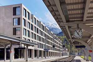 Una estación de tren moderna con un reloj, andén y un paisaje montañoso al fondo.