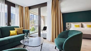 A modern hotel room with a green sofa, a teal armchair, a small table with bottles, and a bed with yellow pillows.