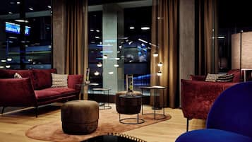 Un salon moderne avec des canapés en velours rouge, un pouf rond et un fauteuil bleu. On y trouve de petites tables avec des lampes, ainsi qu’une grande fenêtre avec des rideaux.
