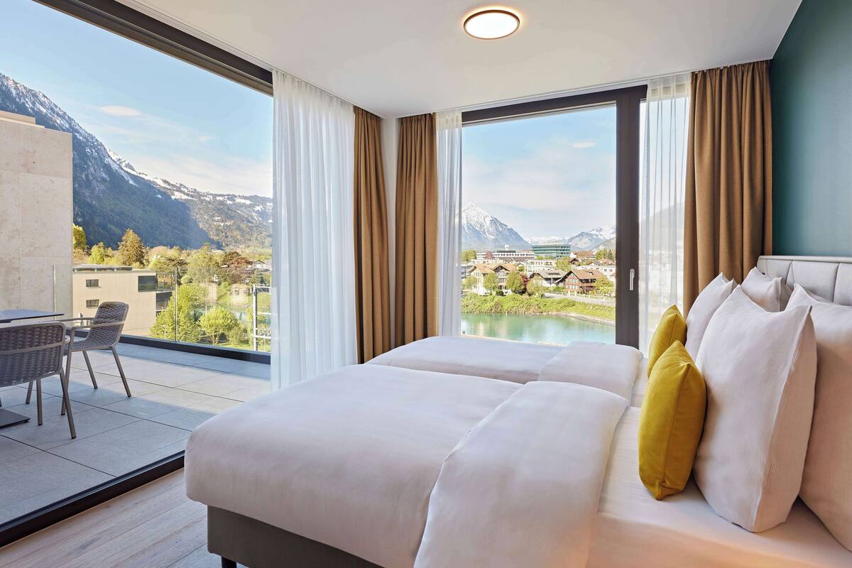 A hotel room with a large bed, a desk with chairs, and a view of mountains and a lake.