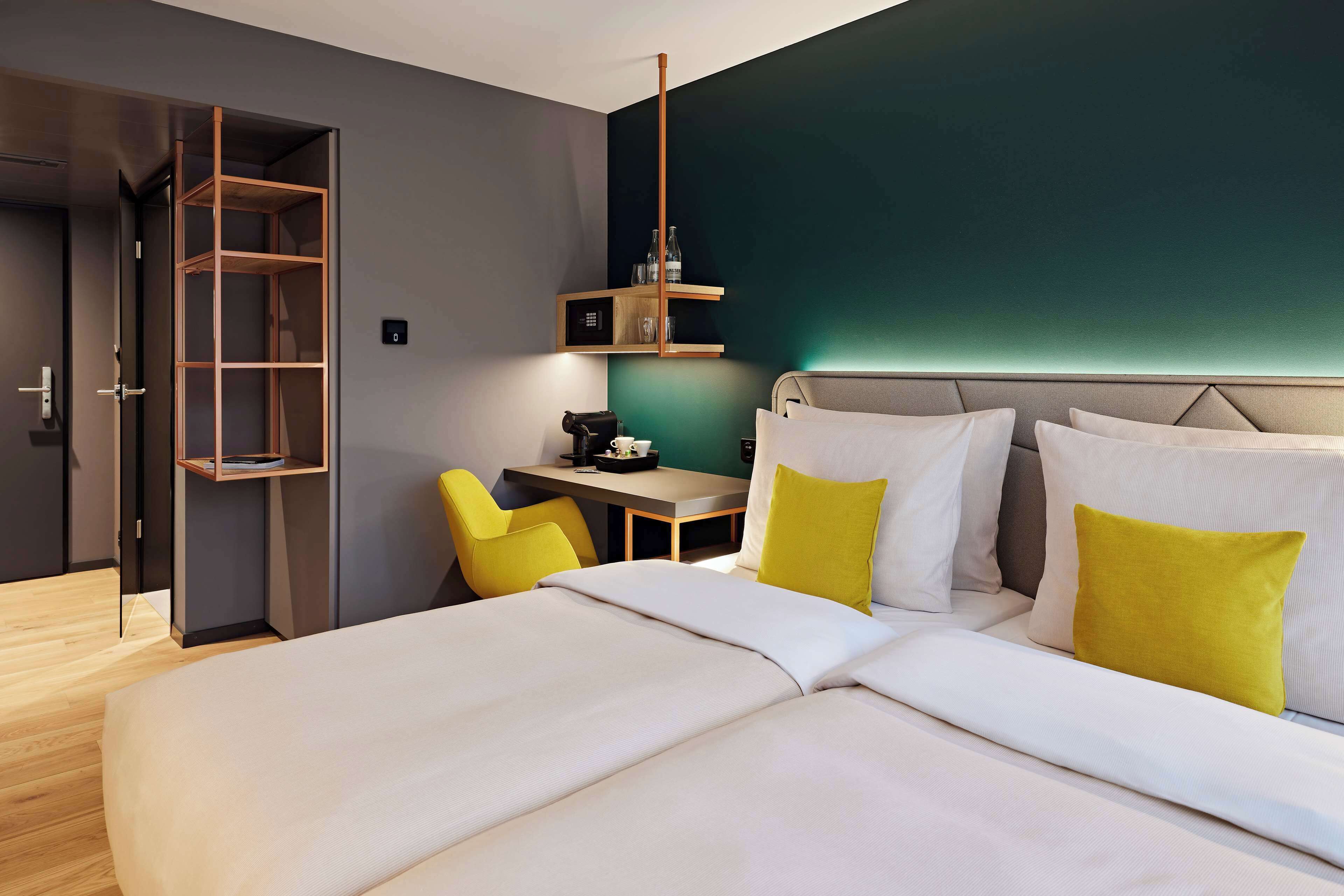 Habitación de hotel moderna con una cama grande, un escritorio y una silla. Hay un armario con espejo y una estantería sobre el escritorio.