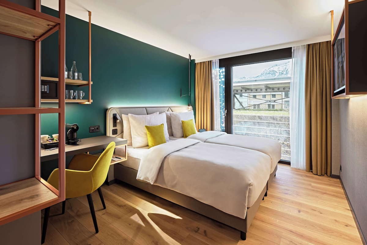 A hotel room with a bed, a desk, a chair, a shelf with glasses, and a view of a bridge.