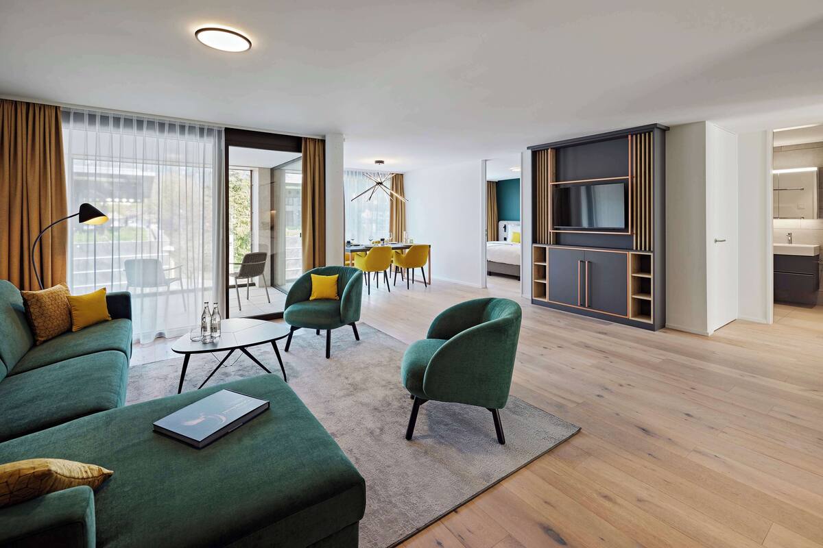 A modern hotel room with a green sofa, yellow chairs, a dining table, and a TV cabinet.
