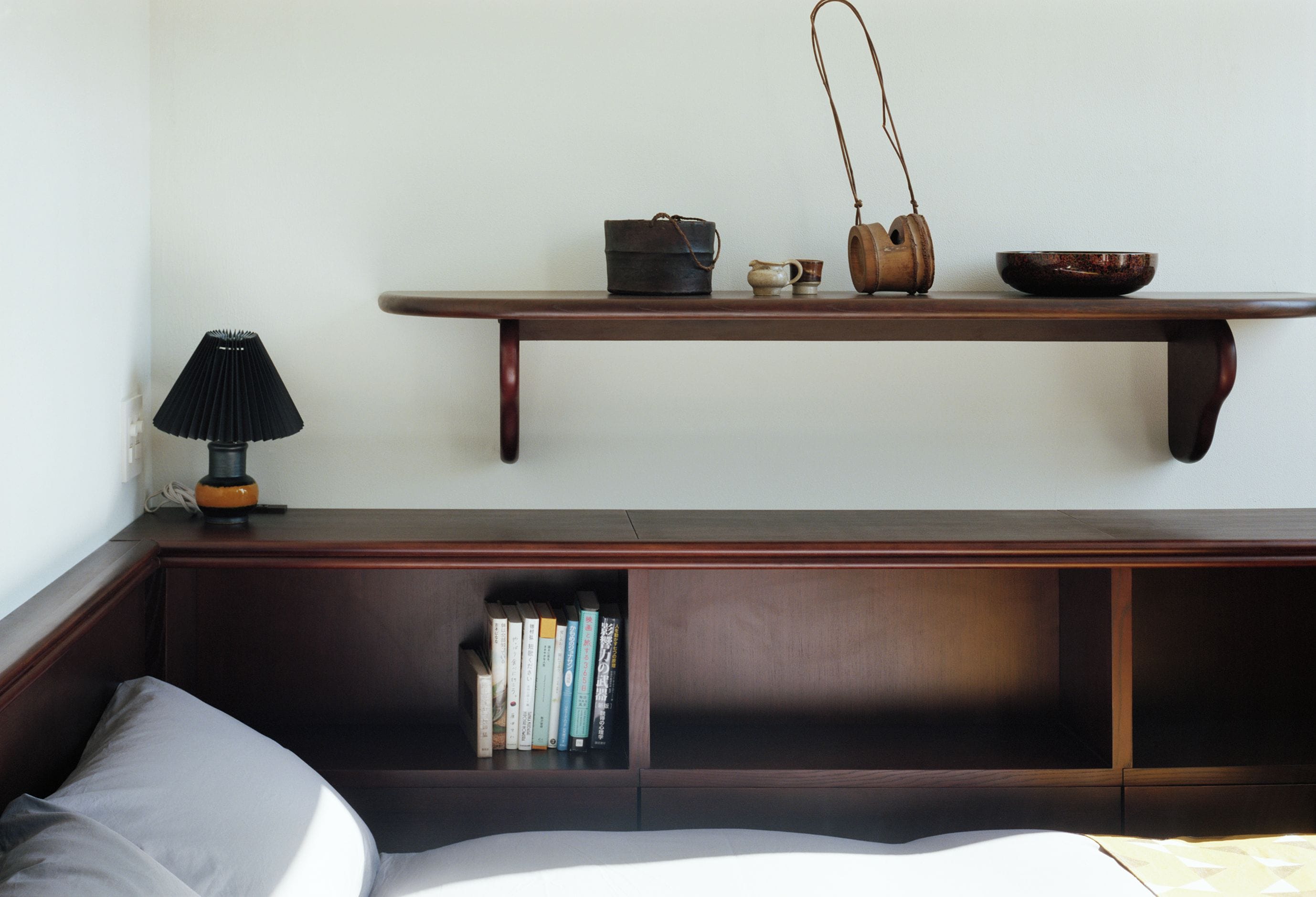 Une chambre avec un lit, une table de chevet avec une lampe, une étagère avec des livres et une étagère suspendue avec un sac et un bol.