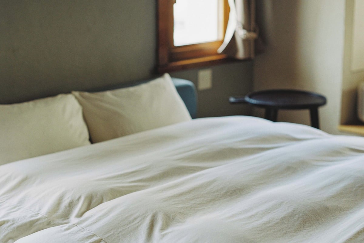 A neatly made bed with white linens in a room with a window and a small round table.