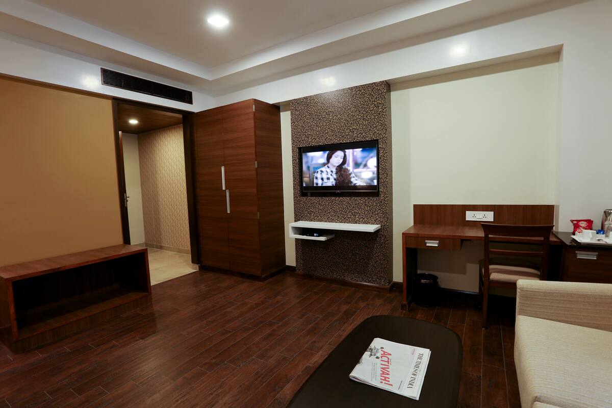 A modern hotel room with a wooden floor, a flat-screen TV mounted on a textured wall, a desk with a chair, a sofa, and a bench.