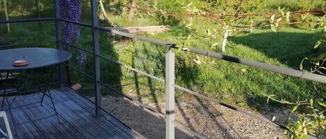 Eine Holzterrasse mit Tisch und Stühlen, umgeben von einem Metallzaun und Blick auf eine Rasenfläche mit Bäumen.