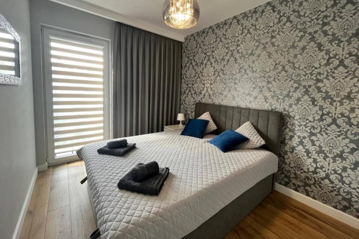 Un dormitorio con una cama grande, papel tapiz gris y blanco con diseño, un cabecero gris y una lámpara colgante.