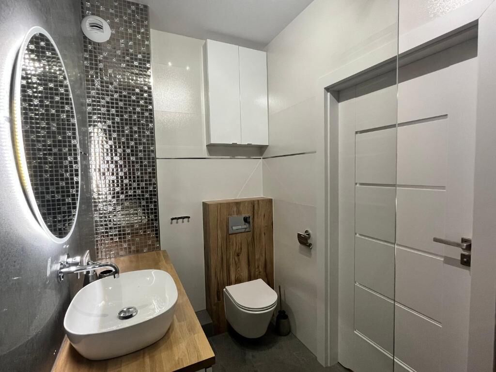 A modern bathroom with a large oval mirror, a white sink, a wooden vanity, a toilet, and a white door.