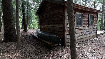Una cabaña rústica en un bosque, con fachada de madera, un porche y una ventana pequeña.