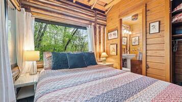 Un dormitorio de cabaña de madera con cama, ventana con cortinas, mesita de noche, lavamanos montado en la pared y cuadros enmarcados.
