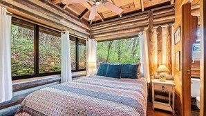 Een slaapkamer in een blokhut met een bed, een nachtkastje, een plafondventilator en grote ramen.