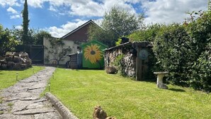 Ein Steinweg führt zu einem rustikalen Gebäude mit einem Sonnenblumengemälde, umgeben von üppiger Vegetation und einer Steinmauer.