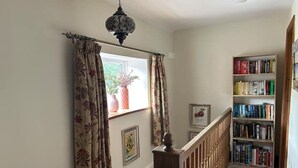 Eine Treppe führt zu einem mit Büchern gefüllten Raum, der ein Holgitter, eine Hängeleuchte und ein mit Vorhängen drapiertes Fenster aufweist.
