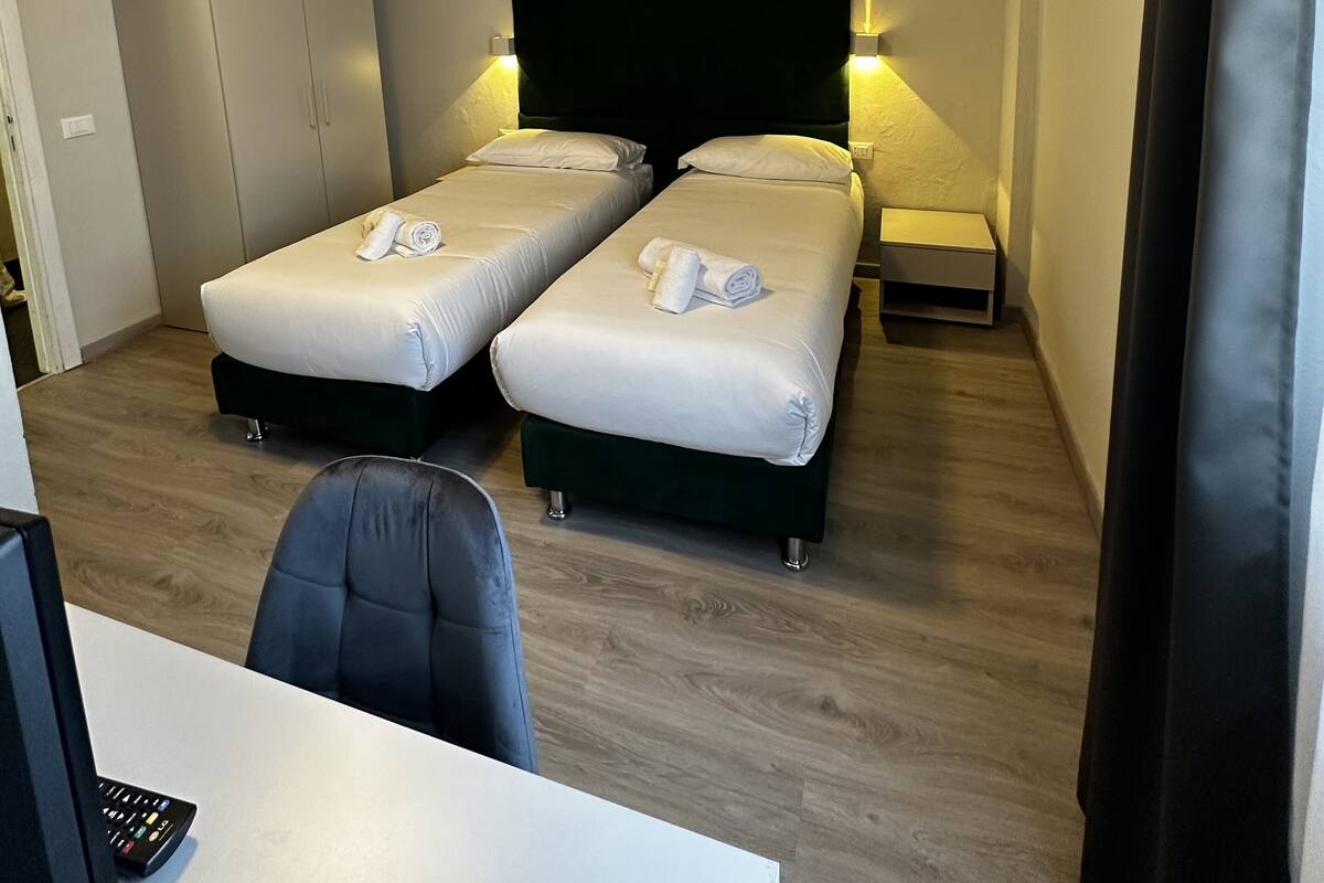 A hotel room with two single beds, a desk with a chair, a flat-screen TV, and a ceiling light.