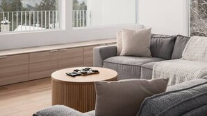 En moderne stue med en grå sofa, et rundt træbord og store vinduer med udsigt til en balkon og bjerge.