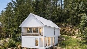 En toetages hytte med balkon, beliggende på en klippefyldt skråning med skov i baggrunden.