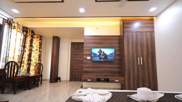 A hotel room with a bed, a TV mounted on the wall, a dining area with chairs, and a wooden wardrobe. - The Crown Hotel (Agra)