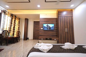 A hotel room with a bed, a TV mounted on the wall, a dining area with chairs, and a wooden wardrobe. - The Crown Hotel (Agra)