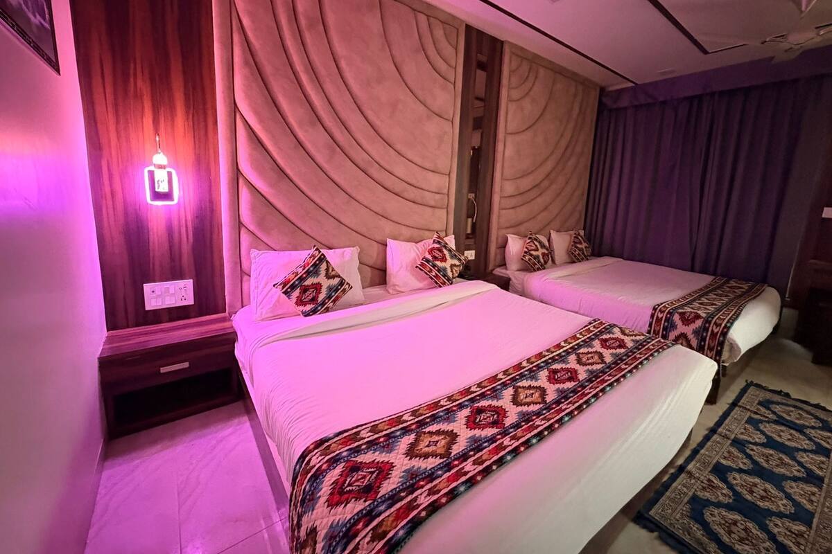 A hotel room with two beds, a wooden headboard, a patterned bedspread, a blue rug, and a wall-mounted light.