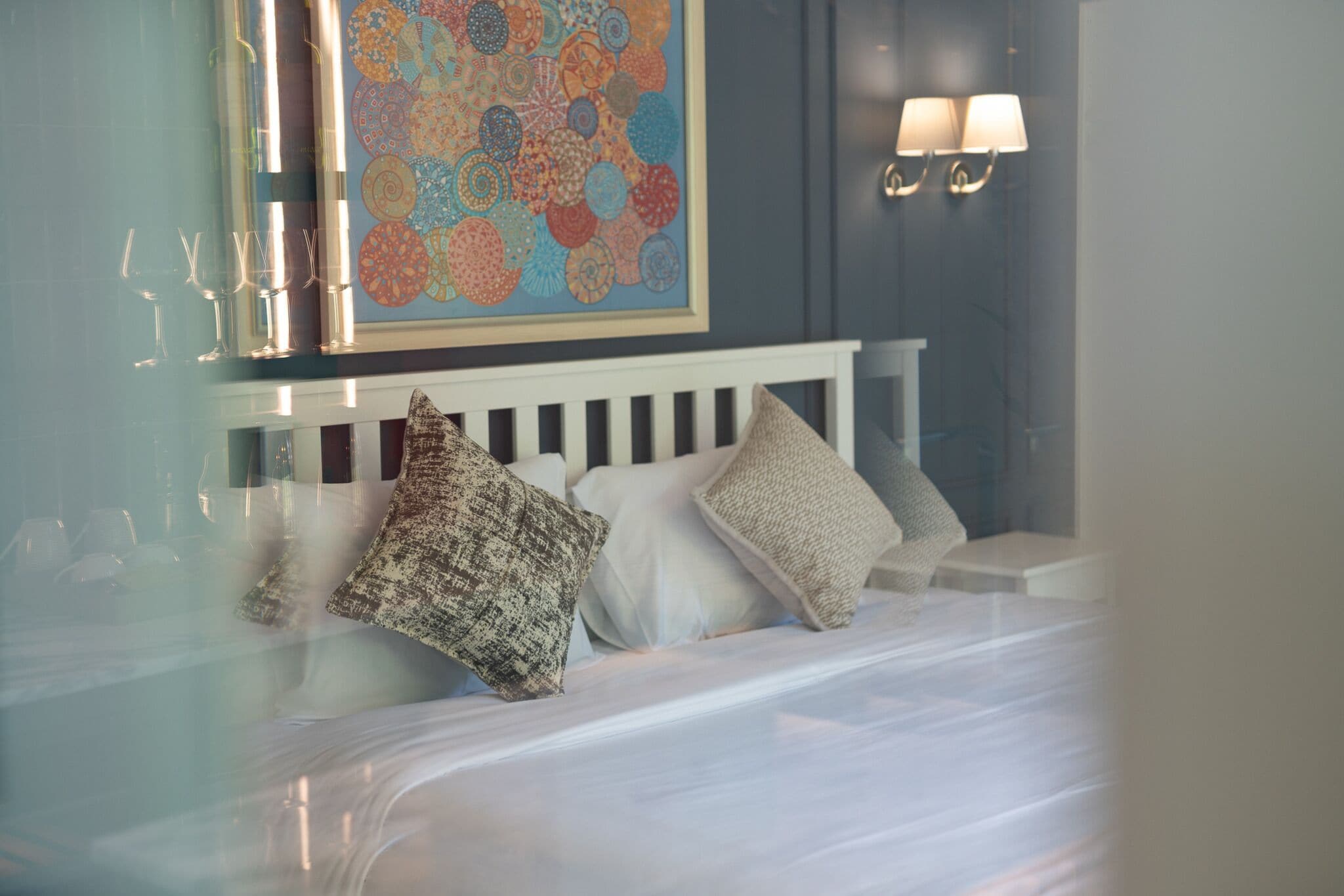 Camera d'albergo con un letto, cuscini, una lampada a parete e un quadro astratto dai colori vivaci.