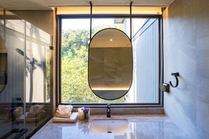 A bathroom with a large oval mirror, a sink, and a window overlooking greenery. - The Stream House at Mae Kampong (Mae On)