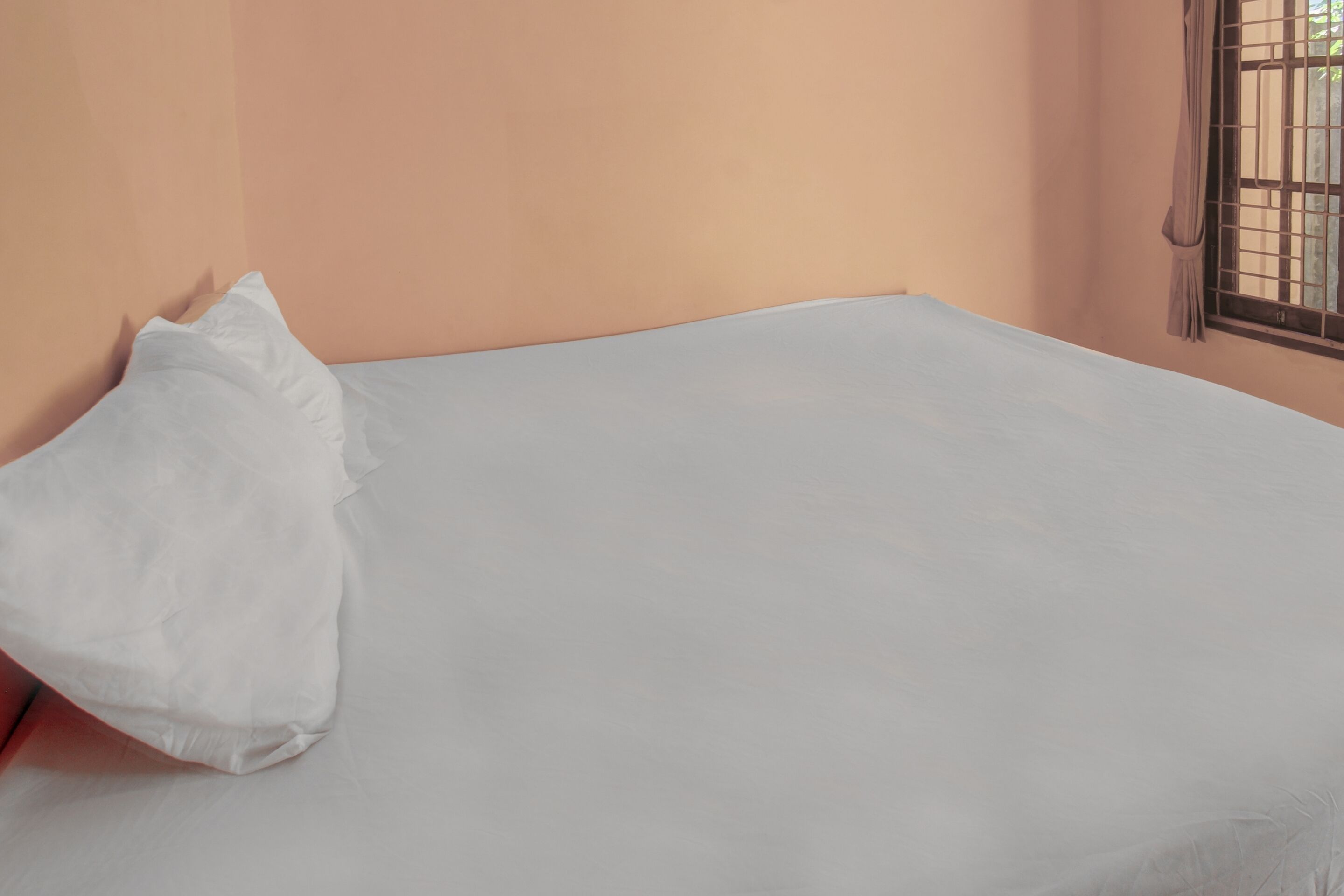 A bed with a white sheet and a single white pillow in a room with peach-colored walls and a window with metal bars.