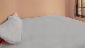 A bed with a white sheet and a single white pillow in a room with peach-colored walls and a window with metal bars.