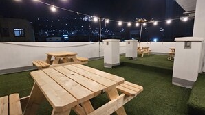 A rooftop terrace with artificial grass, wooden picnic tables, and string lights. - Anjin's Guest House (Mokpo)