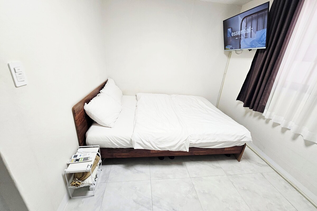 A small, neatly arranged bedroom with a single bed, a TV mounted on the wall, and a bedside table with a remote control.