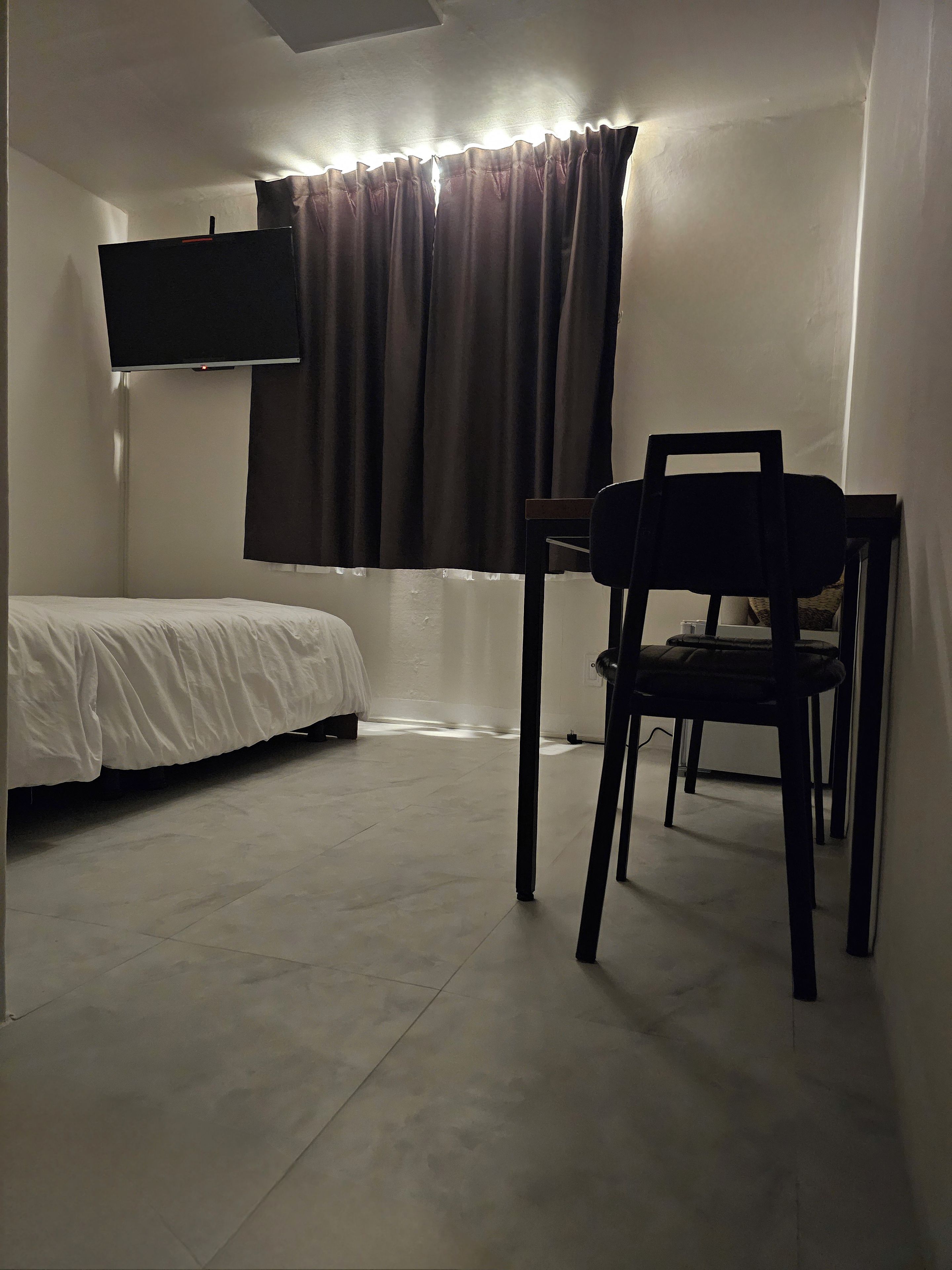 Una habitación con una cama, una silla, un televisor de pantalla plana en la pared y un ventanal con cortinas.