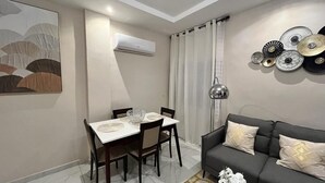 A modern living room with a dining area, featuring a grey sofa, a dining table with chairs, a wall-mounted air conditioner, and decorative plates on the wall. - We Offer you a Lovely 1-bed Apartment in Abidjan (Abidjan)