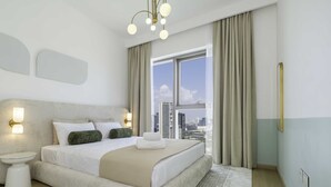 A modern hotel room with a large bed, a bedside table, a wall-mounted light fixture, and a view of the city through large windows. - LUX The Downtown Zabeel View Suite 2 (Dubai)