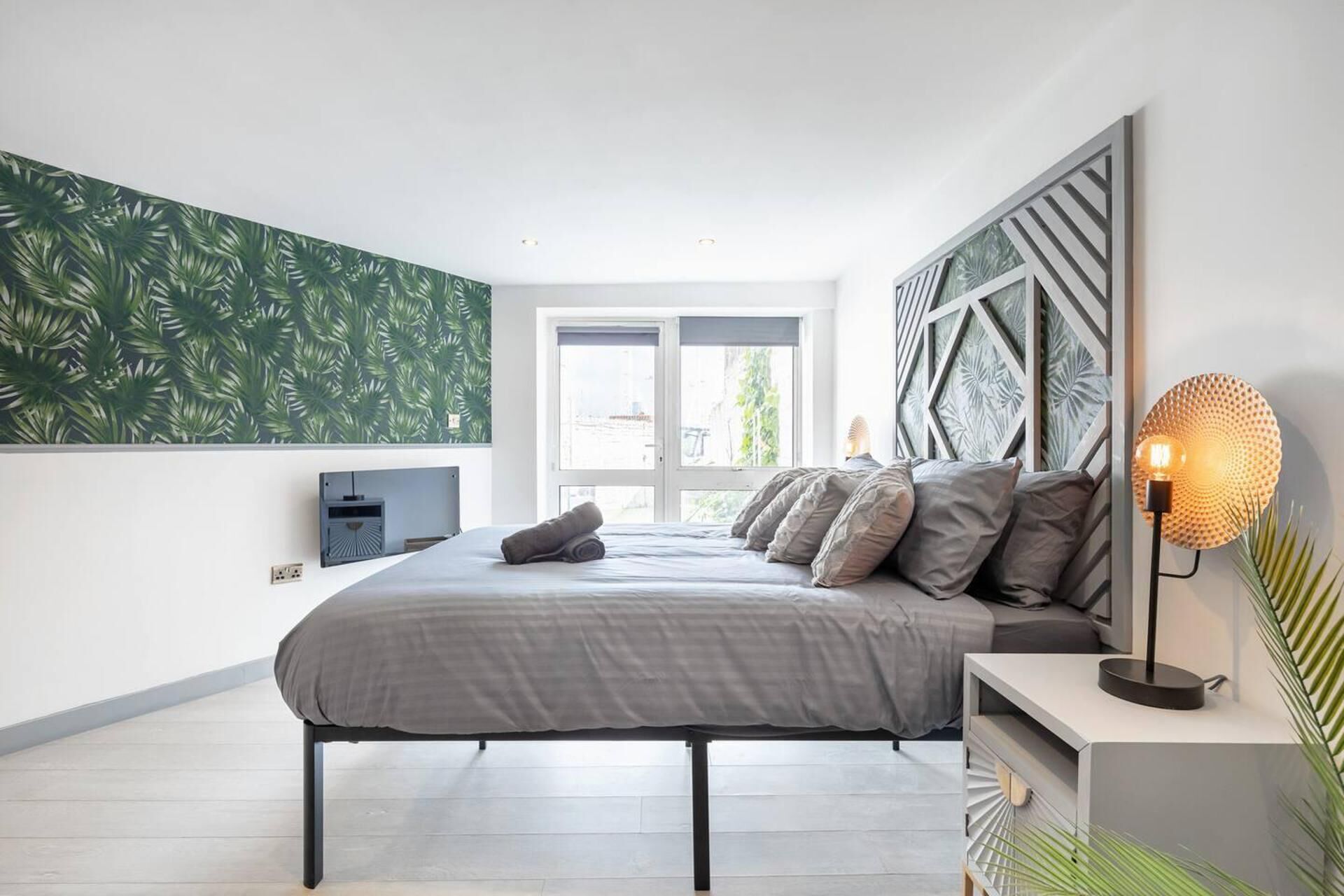 A modern bedroom with a bed, a nightstand, a wall-mounted lamp, and a window with a view.