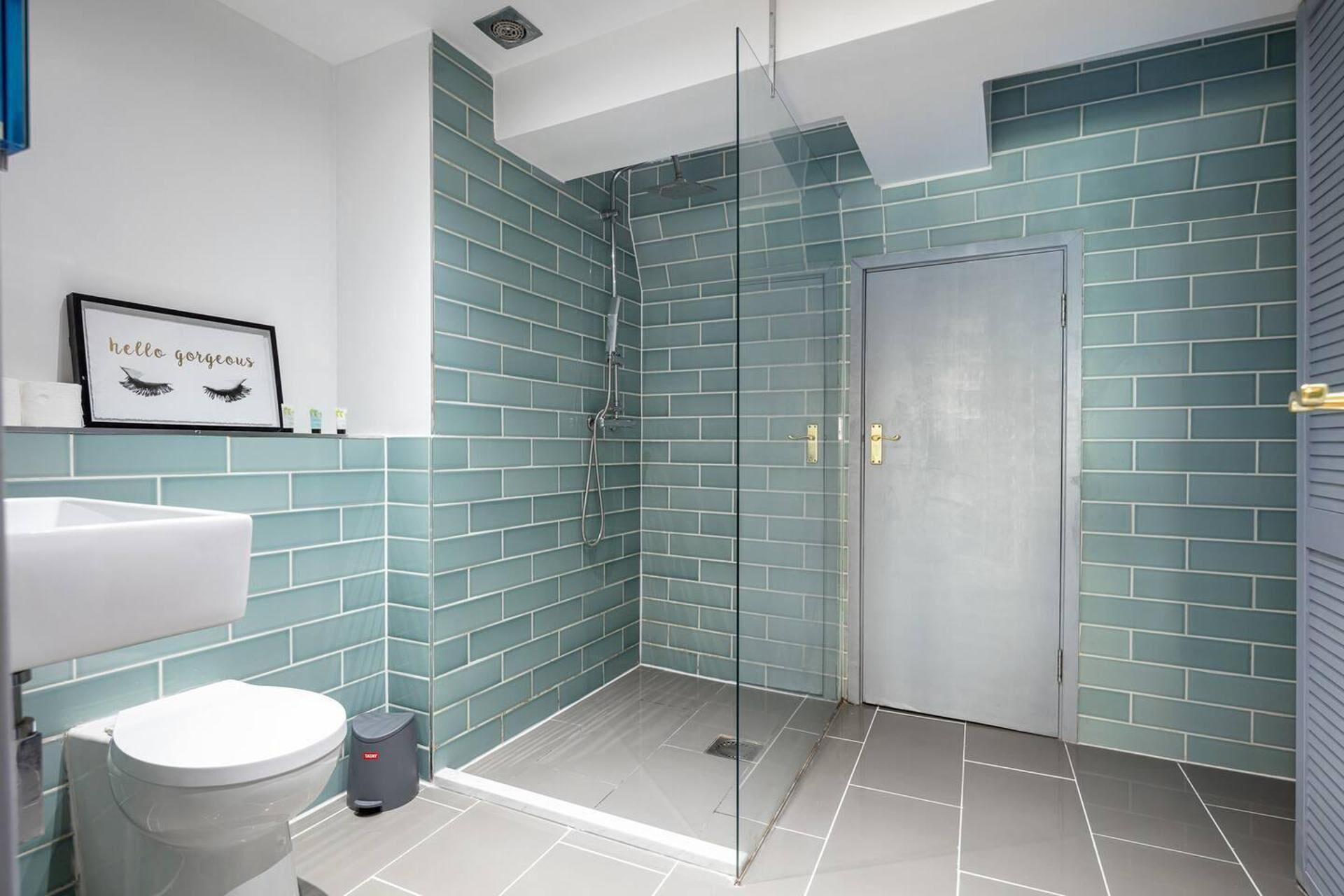 A modern bathroom with teal tiles, a glass shower enclosure, a white sink, and a framed picture on the wall.