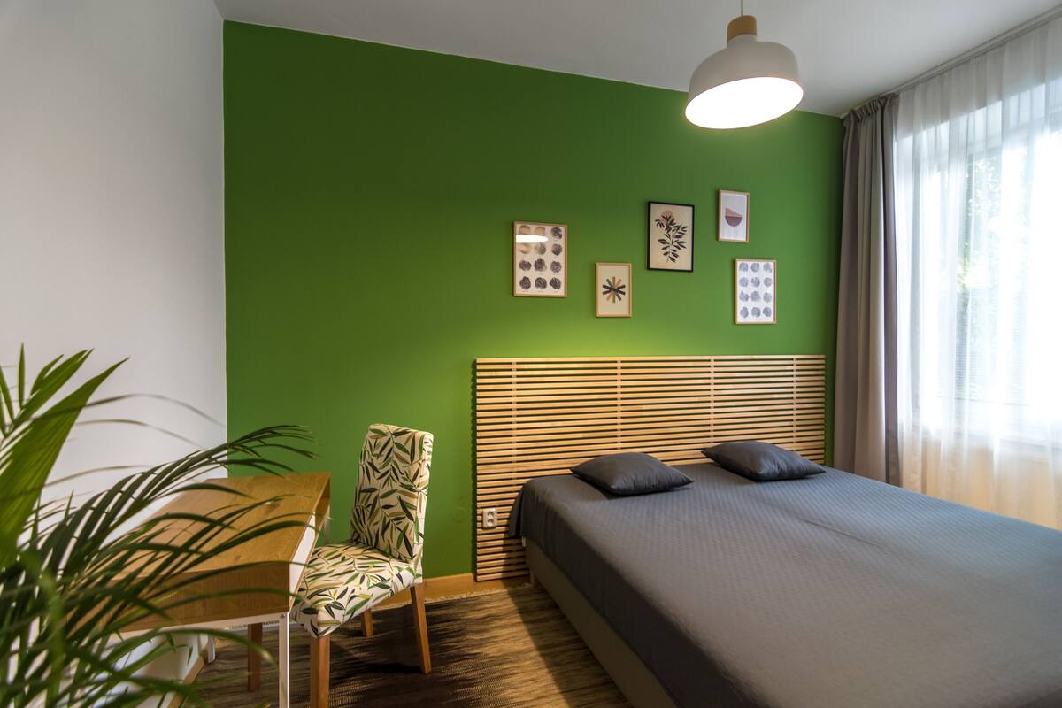 A bedroom with a green wall, a bed with a headboard, a chair, and a table.